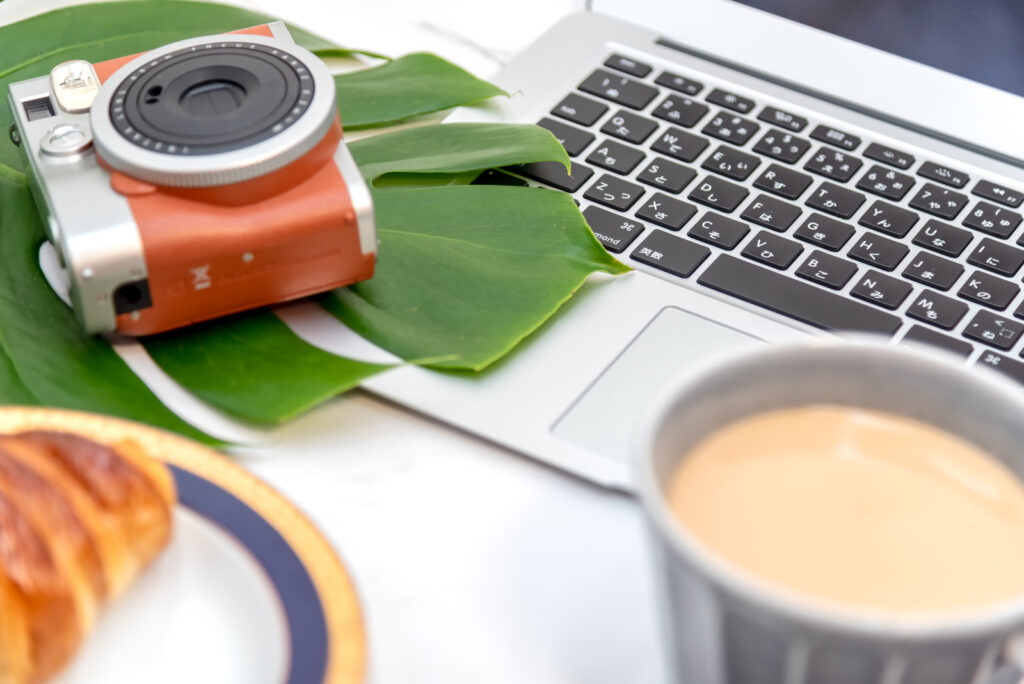 白い机の上にmacbookとカフェオレが入ったカップ、赤いカメラがある。