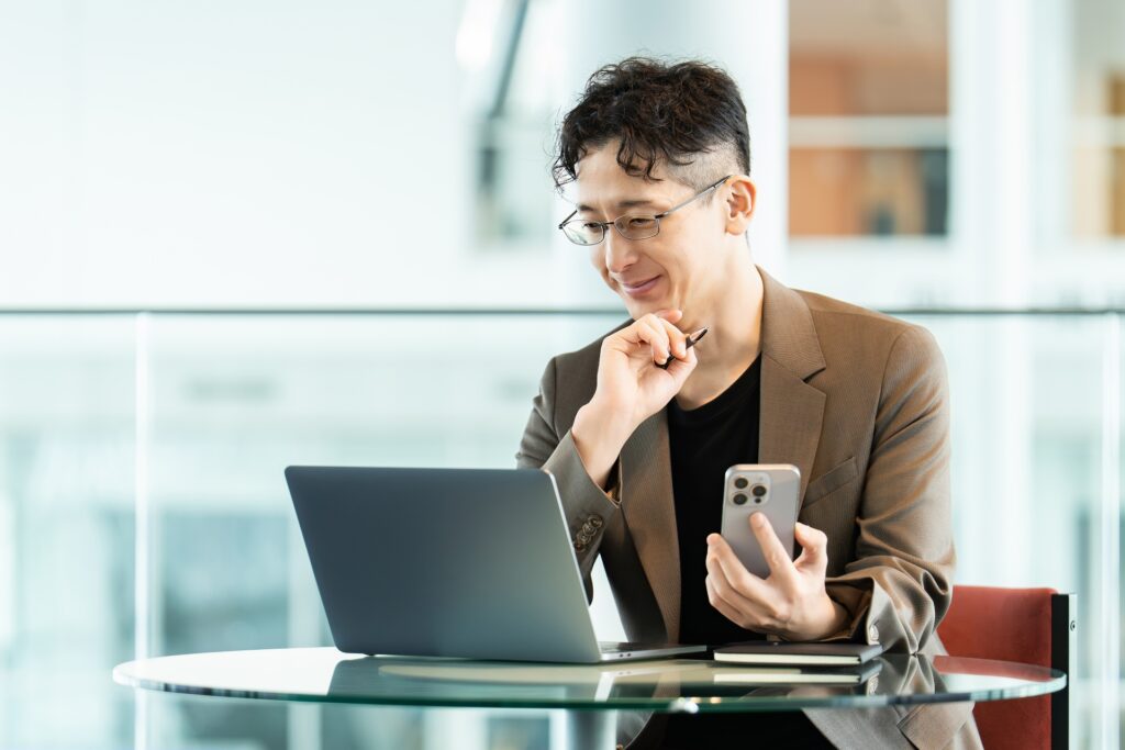 左手にスマホ、右手で頬杖をついてPCに向かっている男性