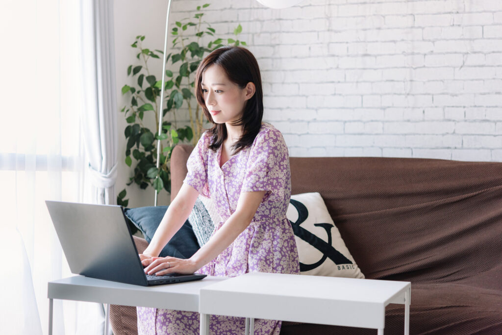 茶色いソファに座ってPCと向かい合っているピンクの花柄のワンピースを着た女性